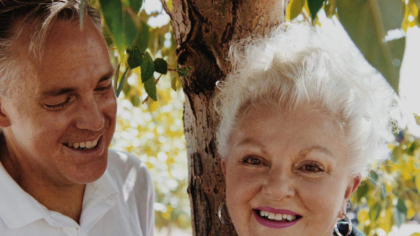 Smiling man and older woman outdoors under a tree